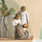 Willow Tree Generations, 7”h hand-painted resin figure of seated older woman sewing blue quilt on her lap; standing woman at her shoulder; seated young girl at her feet, all in neutral clothing, on table with green florals