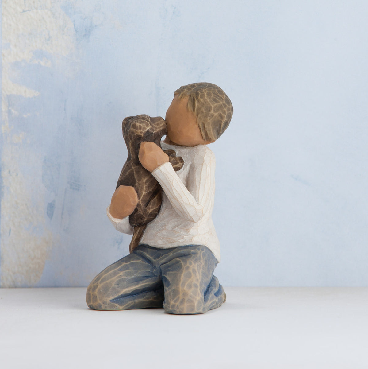 Willow Tree Kindness (boy), 3" hand-painted resin figure in neutral clothes, darker skin and hair, kneeling and holding black dog, in front of pale blue painting