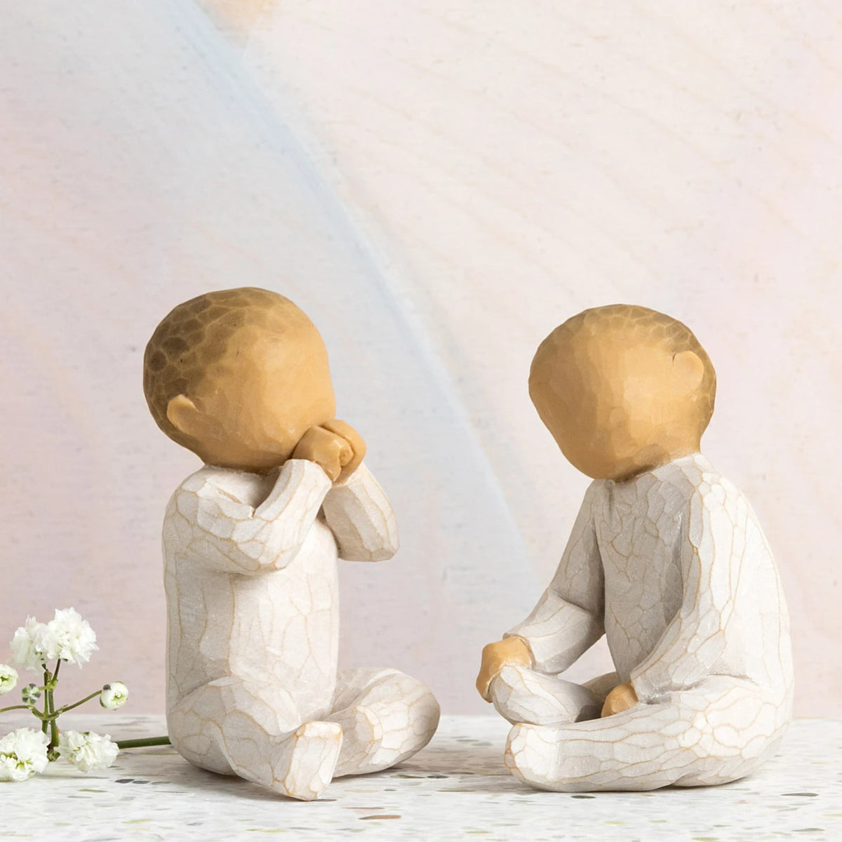 Willow Tree Two Together, two hand-painted seated resin figures of babies, tallest 2", one with hands at mouth, one with hands on legs, both in cream onsies, in front of pastel painting