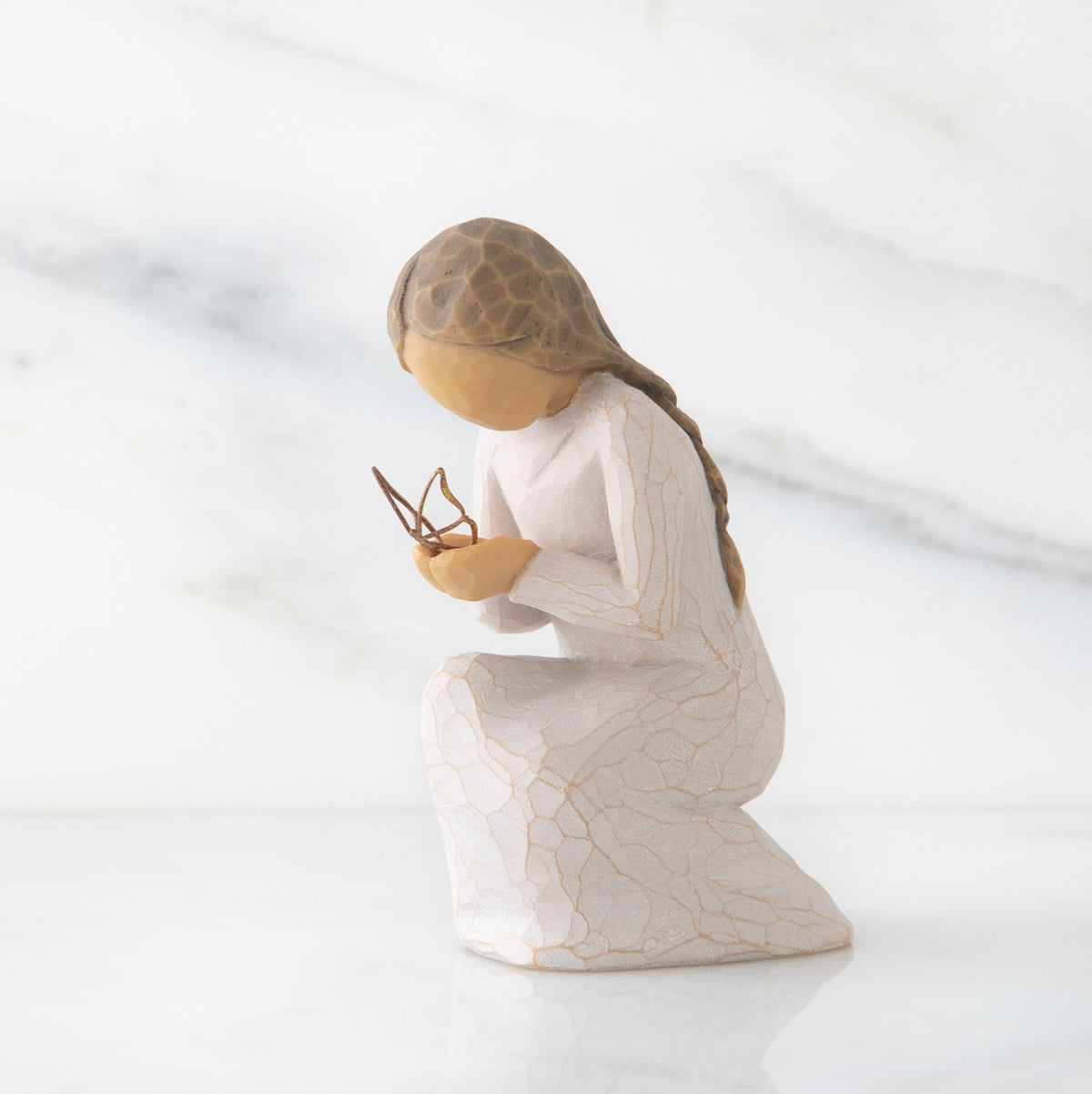 Side view of kneeling female figure in cream dress, with wire butterfly on extended hands, on marble background