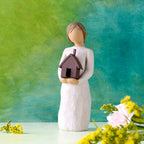 Willow Tree Mi Casa, 5.5”h hand-painted standing resin figure in cream dress, holding dimensional metal house in her arms, in front of deep teal painting