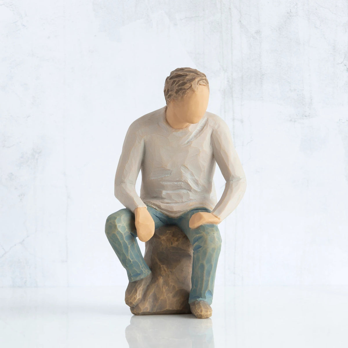 Front view, adult male figure in light gray shirt and blue jeans, seated on gray rock