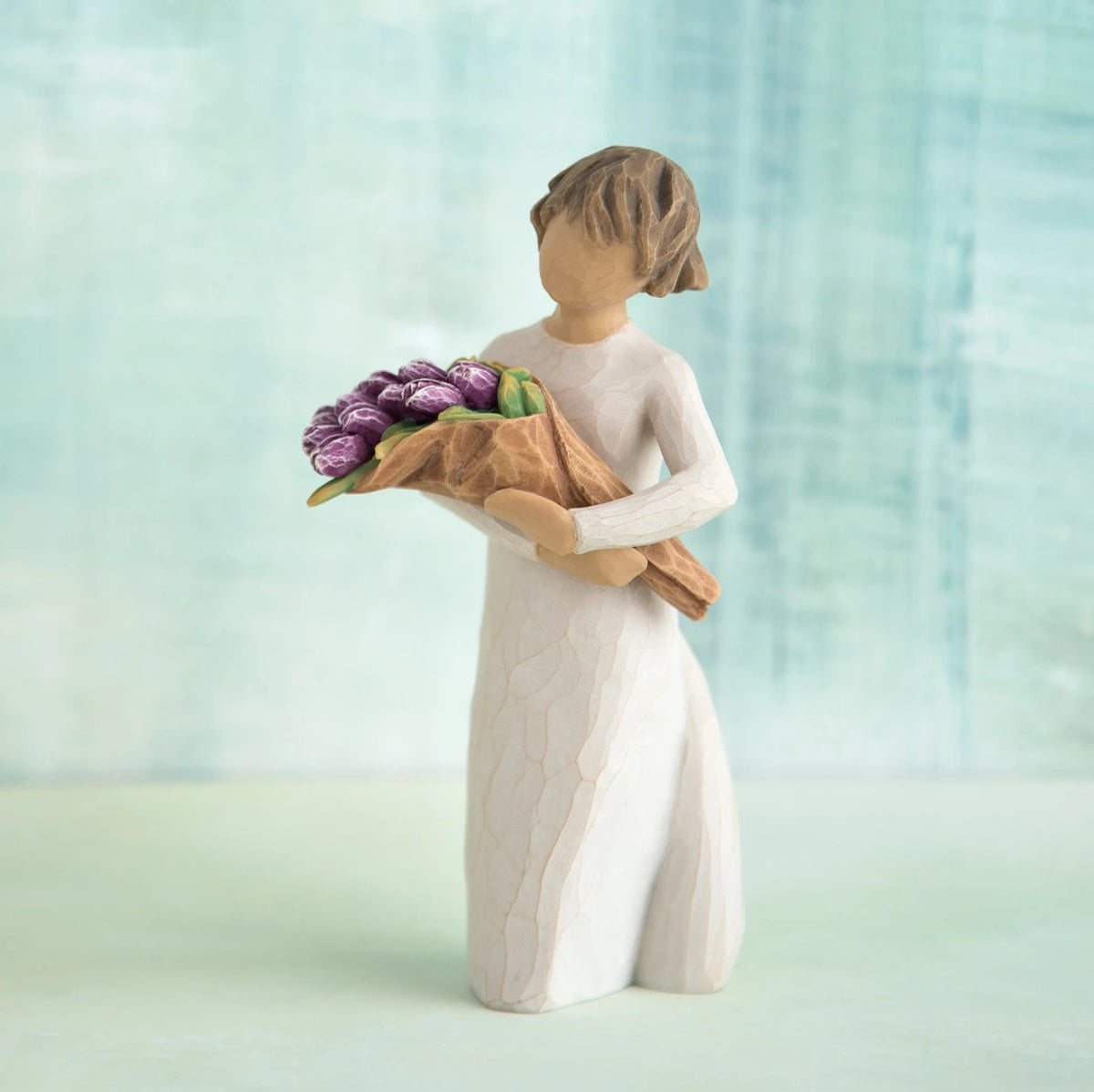 Willow Tree Surprise, 5”h hand-painted standing resin figure in cream dress, holding bouquet of purple tulips, in front of green-blue painting