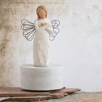 Willow Tree Remembrance Musical, 7.5”h hand-painted resin angel figure in cream dress carved with symbols of nature, wire wings, crossed hands under small gold-leaf heart, atop round, cream base containing musical element, in front of cream painting