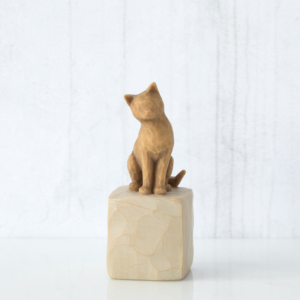 Figure of orange cat with tilted head perched atop a cream colored rectangular pedestal, facing front