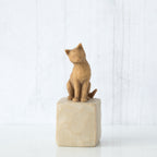 Figure of orange cat with tilted head perched atop a cream colored rectangular pedestal, facing front