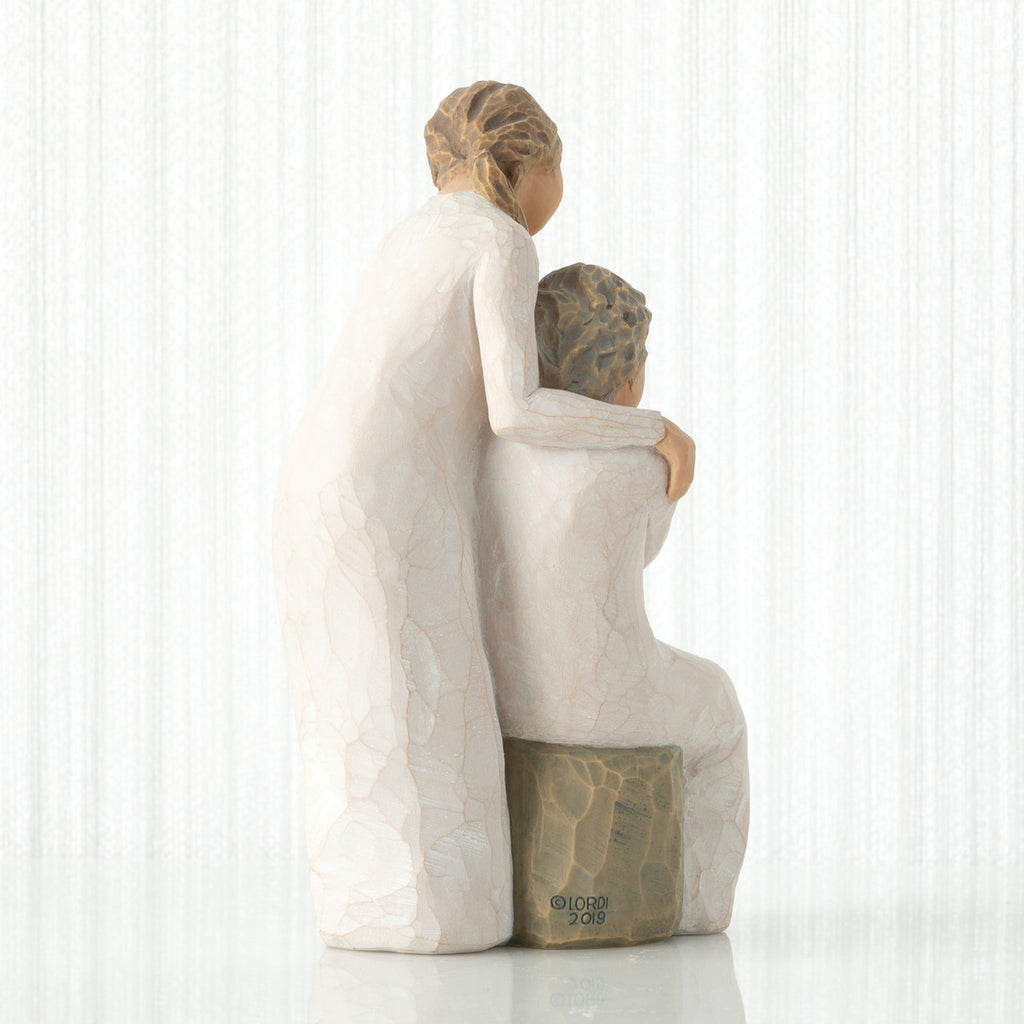 Back view, seated older female in cream dress, with younger female in cream dress, standing at her shoulder, hands embracing