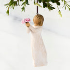 Front view of female figure in cream dress holding out small bouquet of pink peonies, ornament loop affixed to top of head