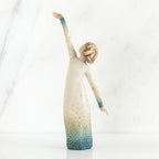 Faceless woman figurine in white dress with blue embellishments - she is facing away from the camera holding both her arms in opposite directions