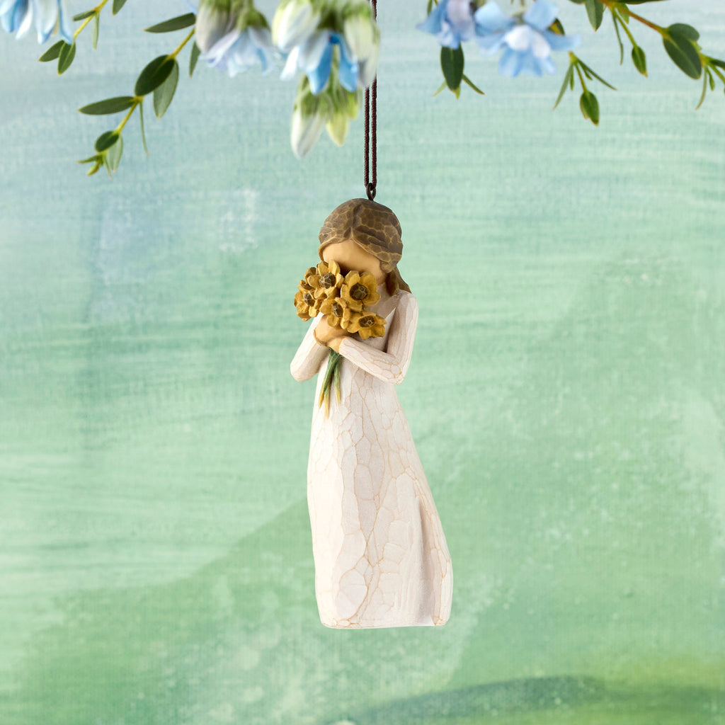 Willow Tree Warm Embrace Ornament, 4.25”h hand-painted resin figure in cream dress, holding bouquet of sunflowers up to face, brown elastic cord, hanging in front of blue green painting