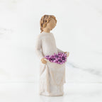 Left side view of woman with tied back, curly, light brown hair holds up a portion of her dress's skirt as it is full of purple flowers