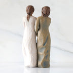 Back view: Figure of two women with darker skin tones in different colored dresses, standing arm in arm