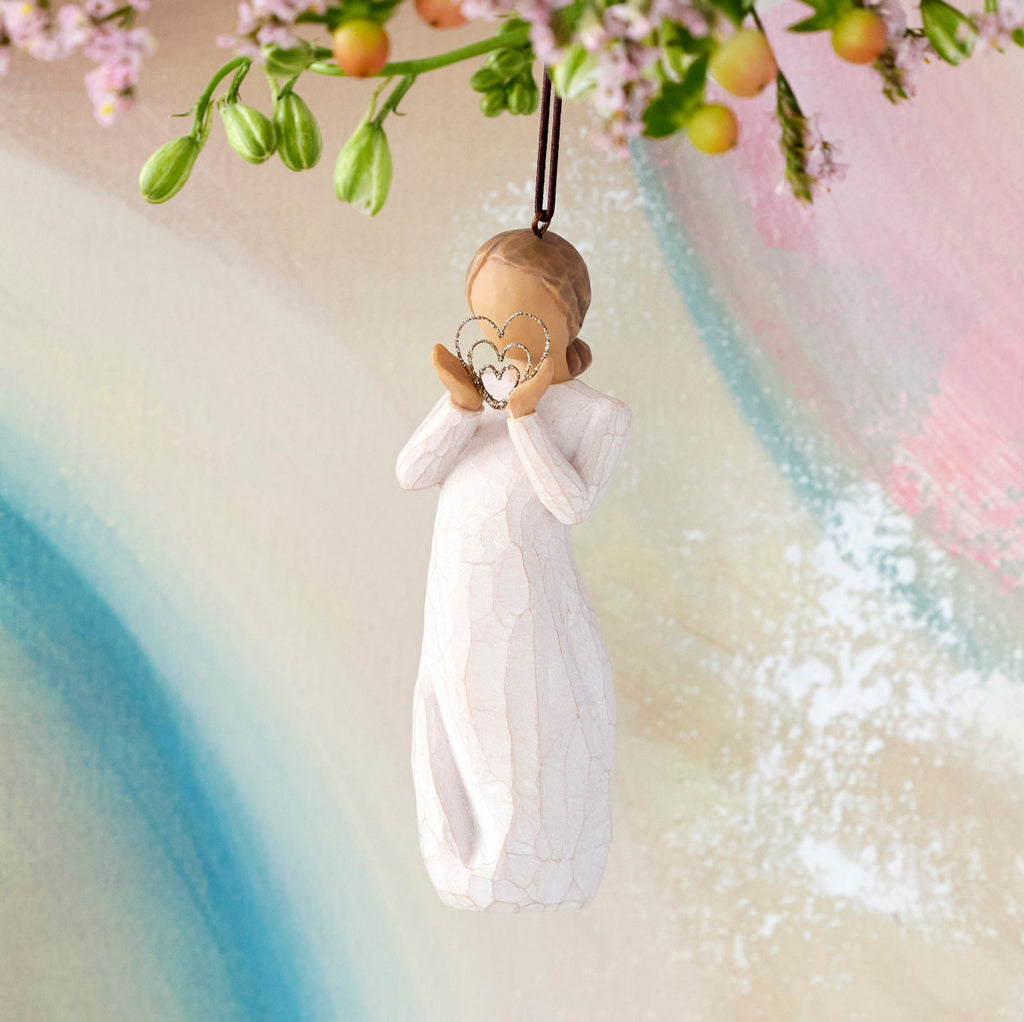 Willow Tree Lots of Love Ornament, 4.5”h hand-painted resin figure in cream dress, holding wire concentric heart with gold glitter, brown elastic cord, hanging in front of multi-pastel painting