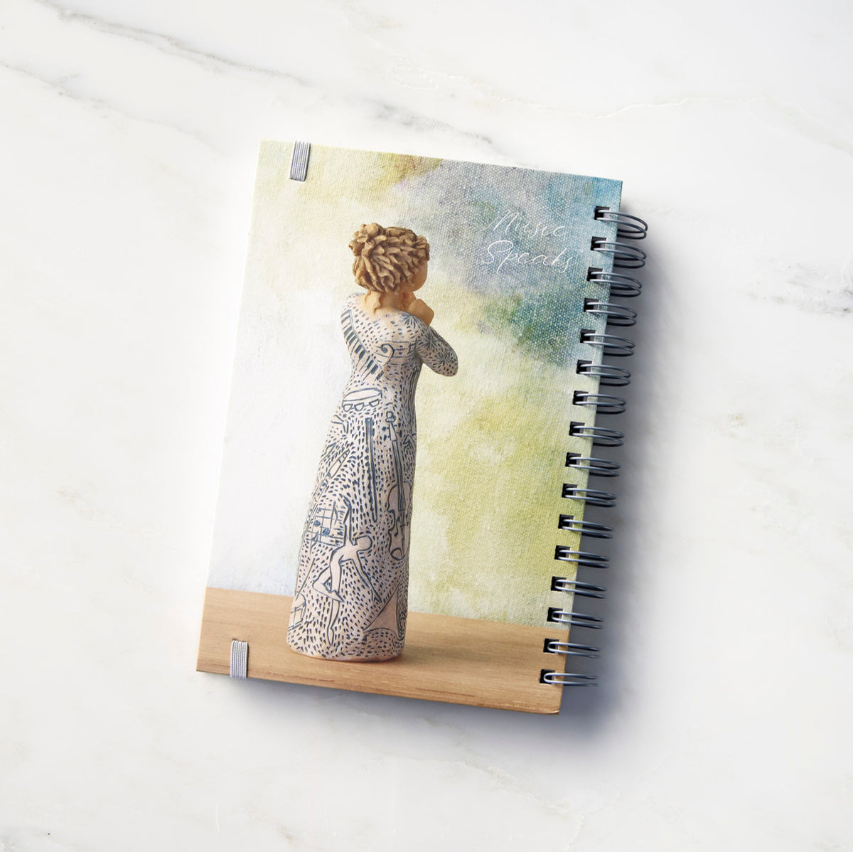 Back view: Spiral-bound notebook featuring back of female figure carved with contrasting pattern of dark blue musical symbols, instruments, dancing forms
