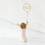 Back view: standing figure in cream dress, arms raised, holding gold wire balloon with 2025 written in gold wire inside
