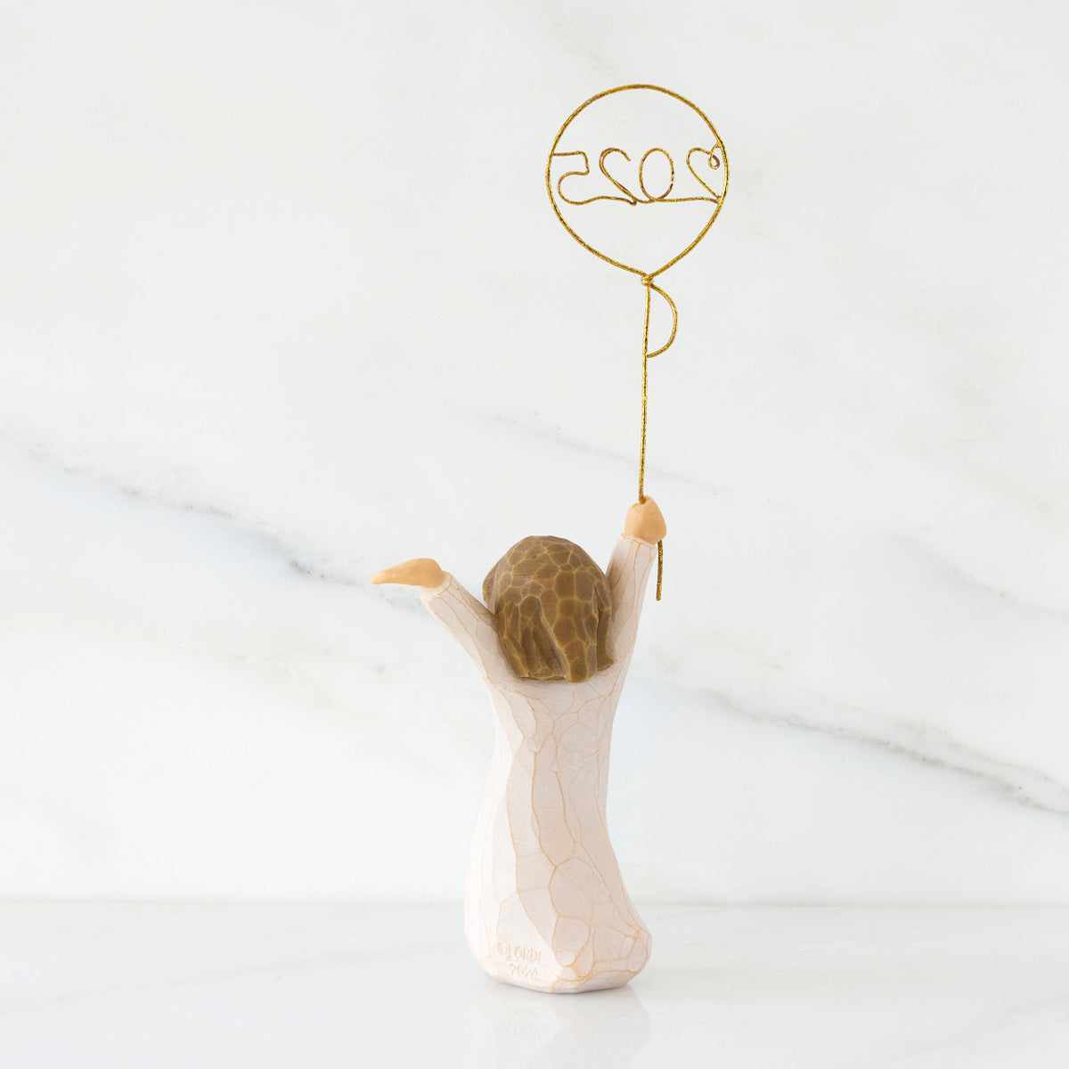 Back view: standing figure in cream dress, arms raised, holding gold wire balloon with 2025 written in gold wire inside