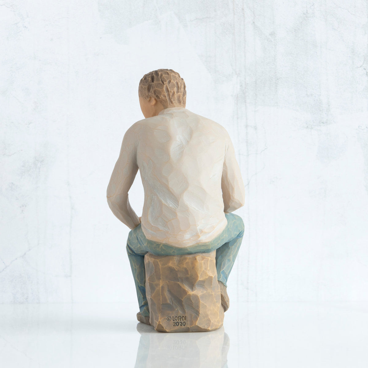 Back view, adult male figure in light gray shirt and blue jeans, seated on gray rock