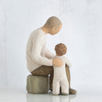 Father figurine in brown pants and white shirt sitting on rock with arm around little boy figurine in white