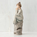 Brunette girl figurine in light grey dress sitting on grey rock facing the side - white background