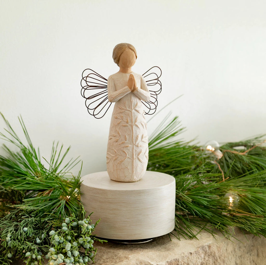 Willow Tree a tree, a prayer Musical, 7.5”h hand-painted resin angel figure in cream dress with wire wings, hands in prayer position, tree carving on dress, atop round, cream base containing musical element, on table with fir branches