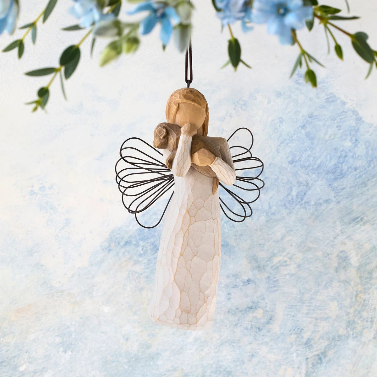 Willow Tree Angel of Friendship Ornament, 4.5"h hand-painted resin figure in cream dress with wire wings holding brown dog, brown elastic cord, hanging in front of sky blue painting
