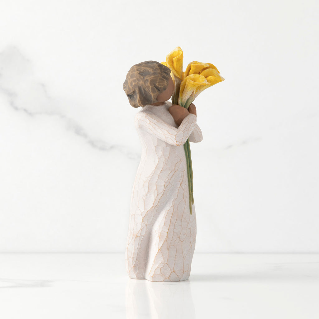 Right side of standing figure in cream dress with darker skin holding yellow calla lilies in her arms