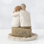 Back view of man and woman figurine in white with arms around another sitting on rock under round white flower plaque