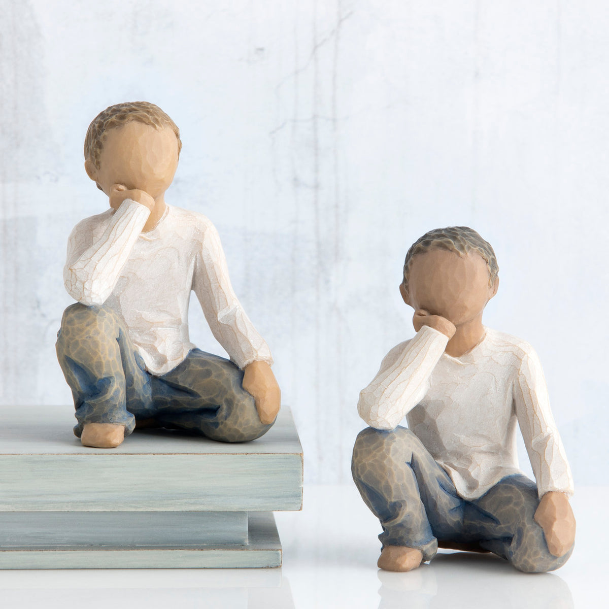 Two young brunette boy figurines in white and jeans sitting down with one arm to their face next to stack of books