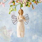 Willow Tree Remembrance Ornament, 4.5”h hand-painted resin angel figure in cream dress carved with symbols of nature, wire wings, crossed hands under small gold-leaf heart, brown elastic cord, hanging in front of sky blue painting