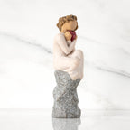 Facing right: figure in cream dress, seated on gray rock, knees drawn up to chest, holding red heart engraved with year 2023