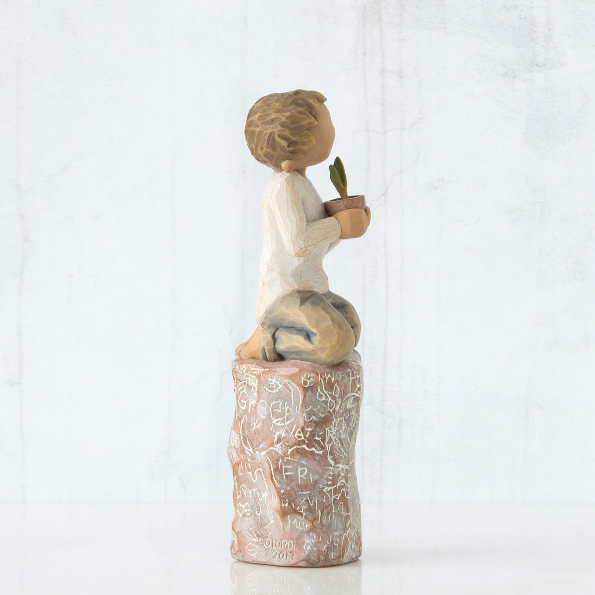 Left side view of boy with brown hair sitting on a stump while holding a small potted plant to his chest