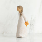 Right side view of girl with brown hair in braid over right shoulder leaning forward as she holds orange flowers behind her back