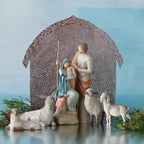 Nativity scene figurines sitting down holding baby next to lambs with brown barn in the background