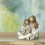Willow Tree Cozy, 3.5”h hand-painted resin female figure sitting with arms around two children, all in cream clothes and wrapped in blue-gray blanket, in front of blue painting