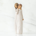 Two faceless woman figurines standing wearing white dresses hugging one another