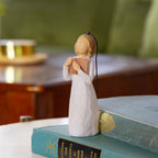 Willow Tree Love of Learning Ornament, 4”h hand-painted resin figure in cream dress holding open book to her chest, brown elastic cord, standing on pile of books