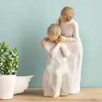 Willow Tree Loving My Mother, 6.5”h hand-painted resin figure of seated older woman with younger woman standing at her shoulder, both in cream dresses, hands clasped, in front of muted teal painting