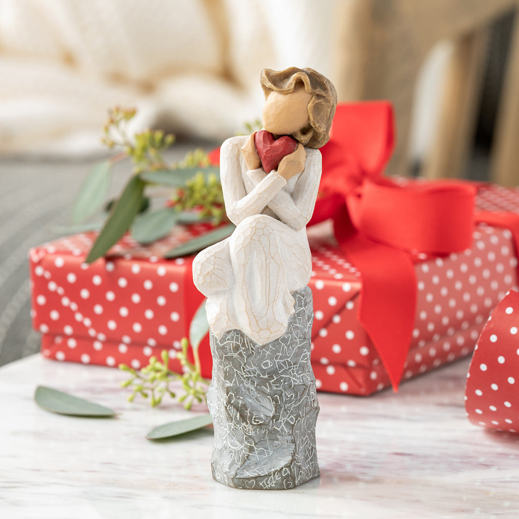 Willow Tree Always, 6”h hand-painted figure in cream dress, knees drawn up to chest, holding red heart to face, seated on gray rock etched with symbols of love, on table with gifts