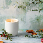 Willow Tree Festive Orange and Frankincense Candle, 3.5"h off-white ceramic vessel with plant-based wax and blend of natural oils, "behold the awe and wonder" sentiment in gold inside rim, in front of blue painting