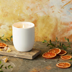 Willow Tree Festive Orange and Frankincense Candle, 3.5"h off-white ceramic vessel with plant-based wax and blend of natural oils, "behold the awe and wonder" sentiment in gold inside rim, in front of orange painting
