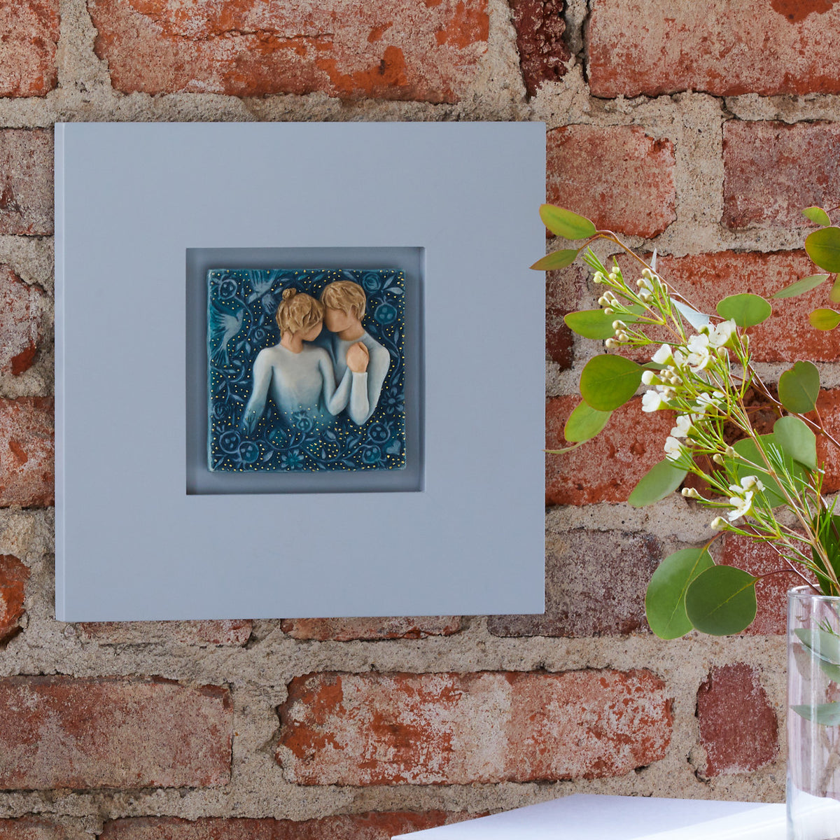Willow Tree Duet, 10”sq hand-painted dove gray wood frame, with inset bas-relief resin plaque of embracing couple, surrounded by nature, blue ombré background with gold leaf, hanging on brick wall